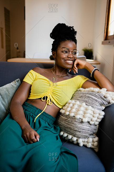 Positive young African American female in bright yellow shirt and transparent eyeglasses looking away with bright friendly toothy smile while relaxing on couch in bright studio apartment