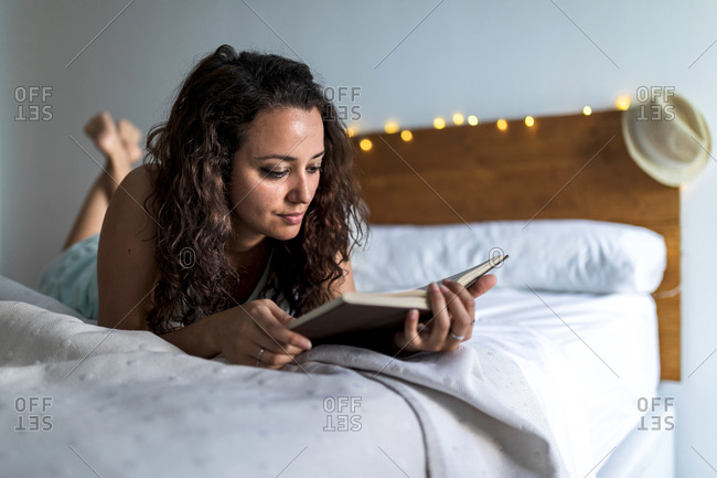 Pensive female lying on comfortable bed and reading interesting story in book while spending weekend at home