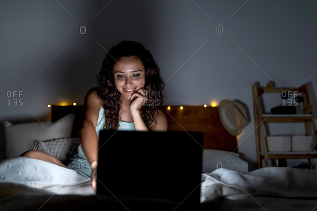 Delighted female in nightwear lying on bed in dark room and watching funny video on netbook while smiling and spending weekend at home