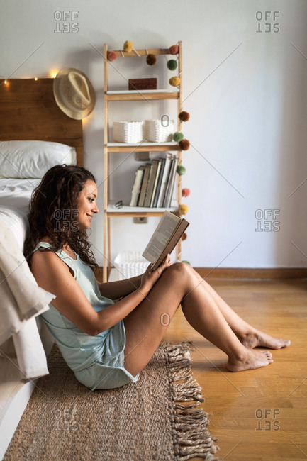 Side view of gentle female in pajamas sitting on floor near bed and enjoying interesting story while smiling and resting at home at weekend