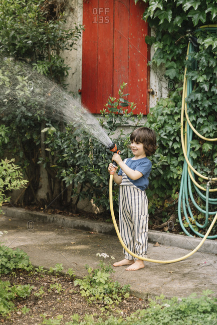 Smiling boy watering plant with garden hose while standing on footpath at back yard
