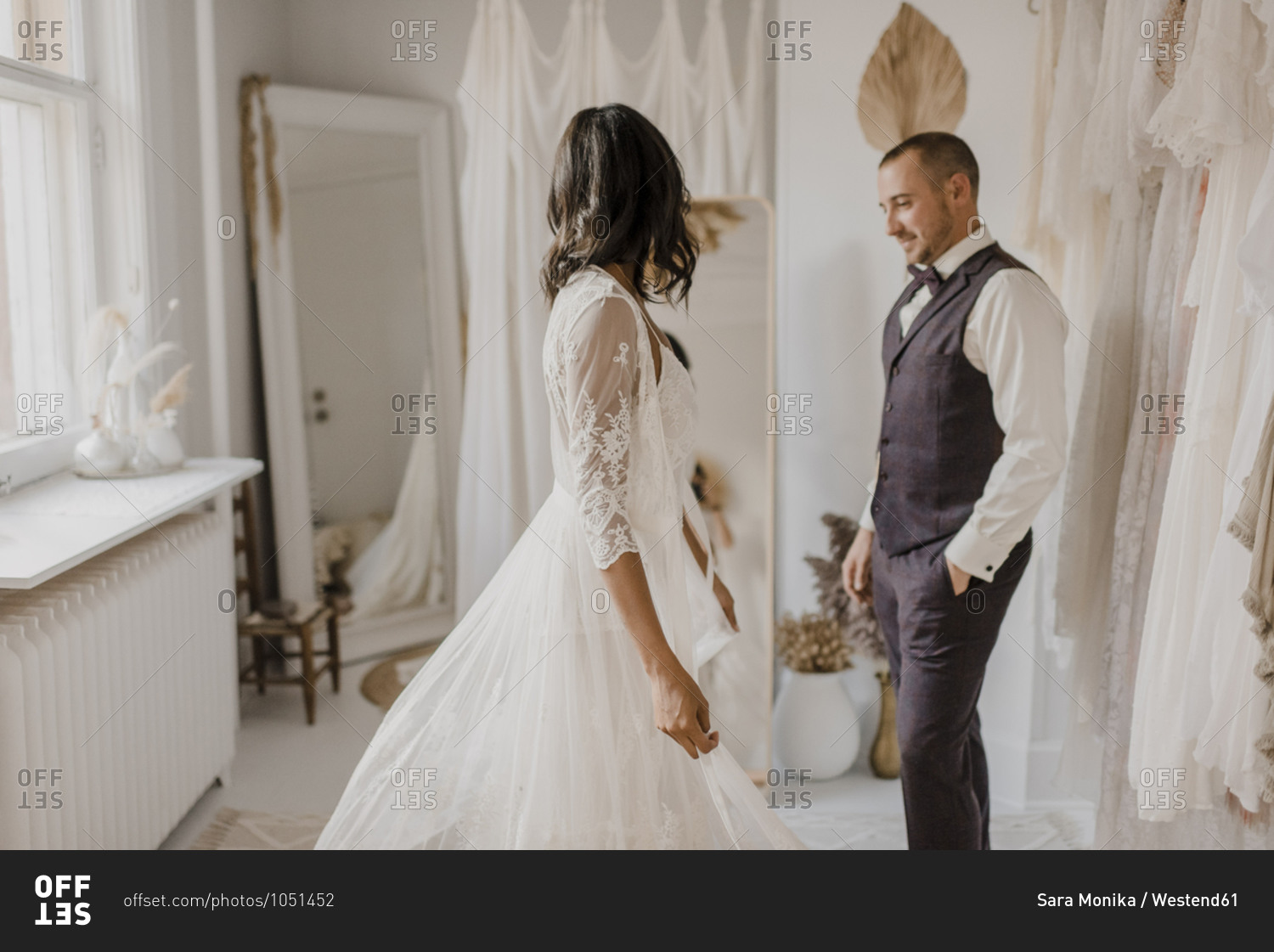 Young bride looking in mirror while bridegroom standing with hands in pocket at home