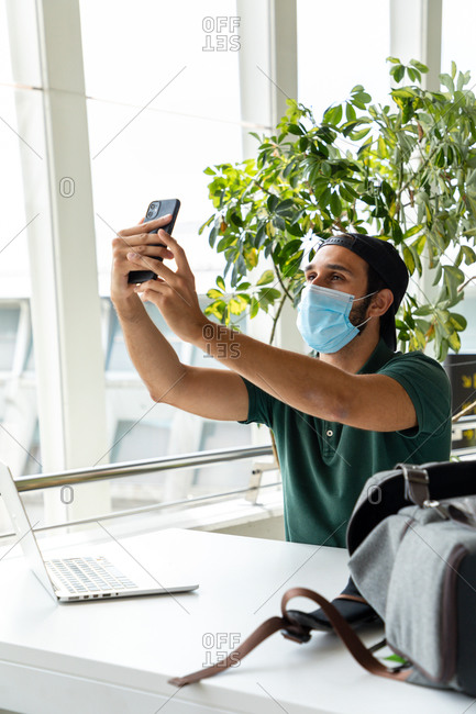 Smiling male wearing medical mask sitting at table with laptop and taking photo on selfie camera of cellphone