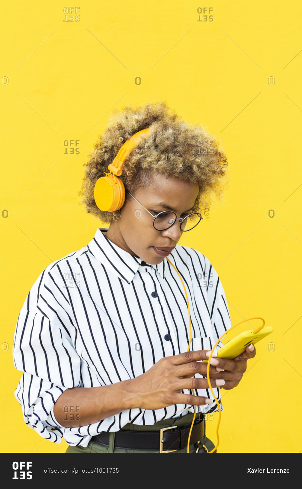 Curly haired woman listing to music while using a smartphone on a yellow background