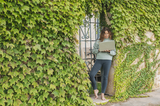 Mature woman using laptop while leaning on ivy wall at gate