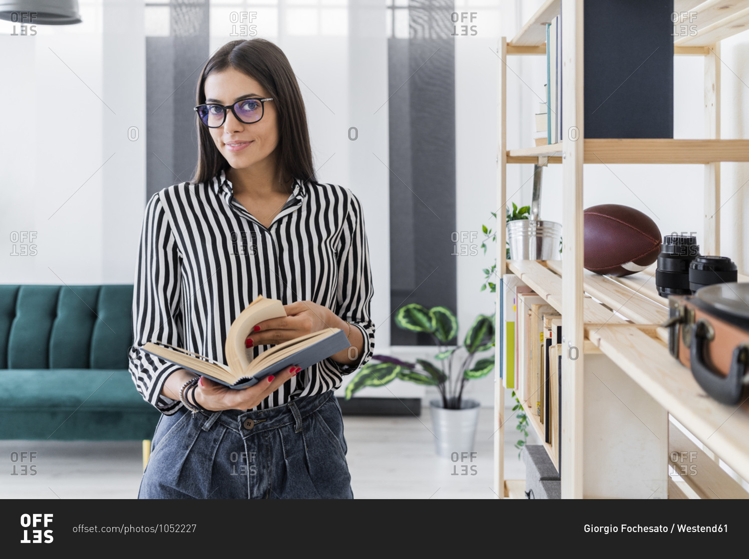Confident beautiful young design professional holding book while standing by rack at creative office
