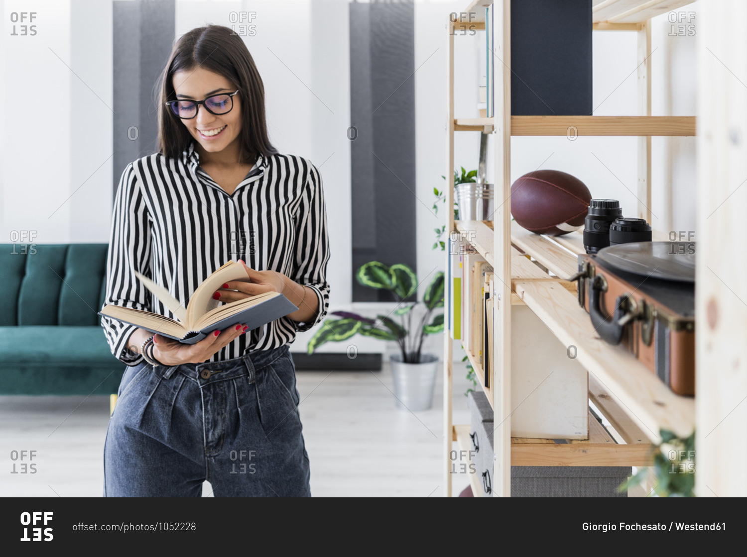 Smiling beautiful young businesswoman reading book while standing by rack at creative office