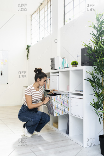 Young businesswoman crouching while searching in box on rack at creative workplace