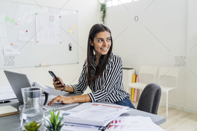 Happy female architect looking away while sitting with wireless technologies at desk in office