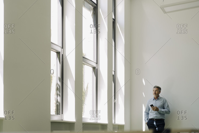 Businessman holding phone while leaning on wall at office