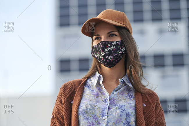 Young woman wearing cap and face mask looking away while standing in city