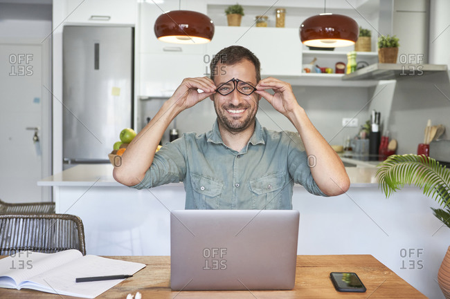 Playful male freelancer holding eyeglasses while sitting with laptop at home
