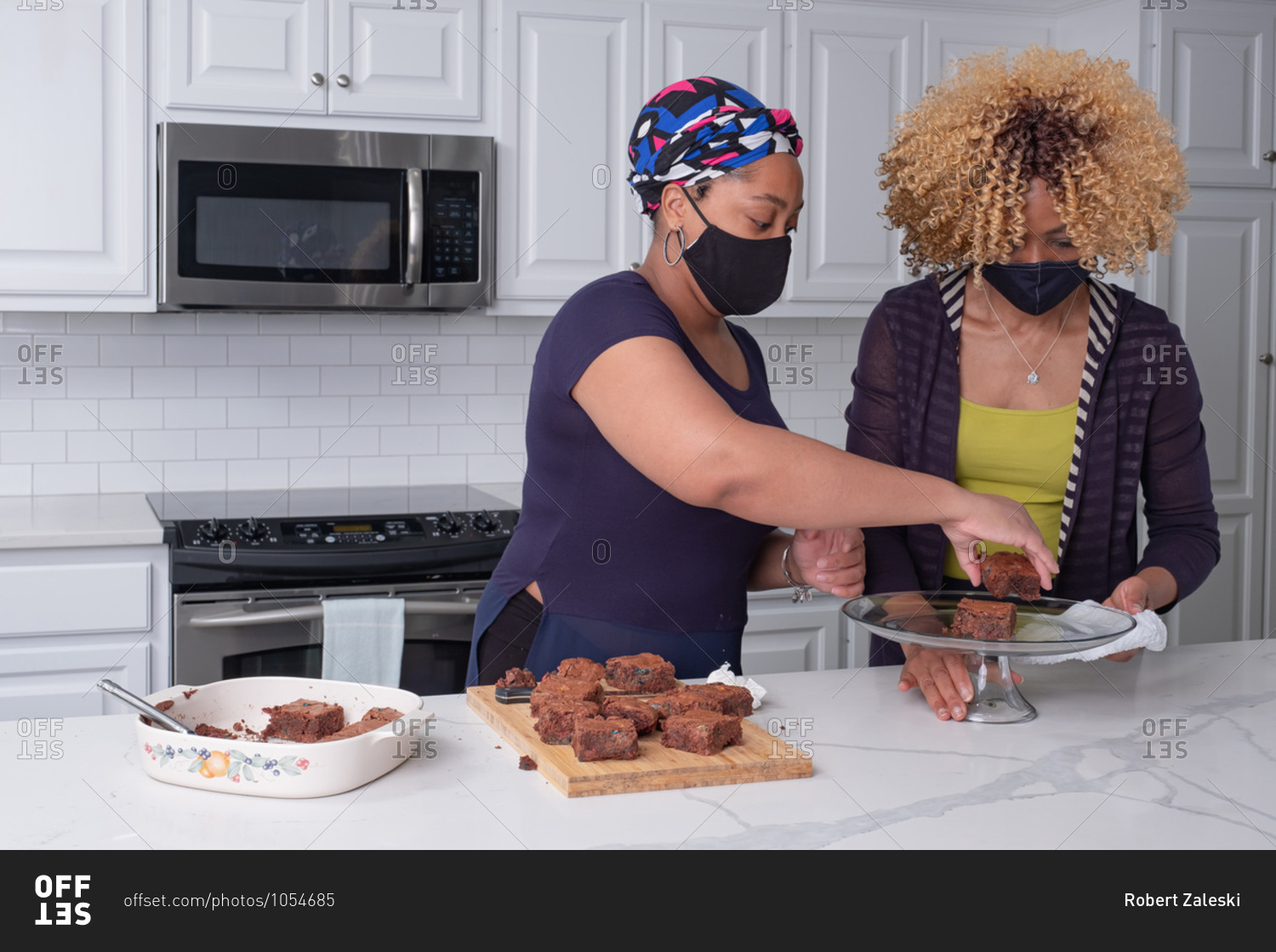 Black mother and daughter arranging brownies on a glass stand while wearing face masks