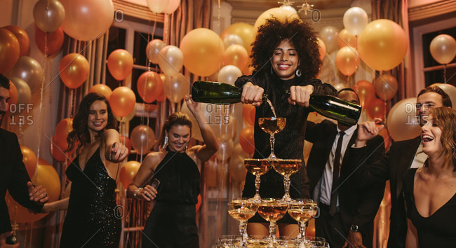 African female filling champagne pyramid with two bottles and friends dancing around at a party. Multi-ethnic group of people enjoying at a party.