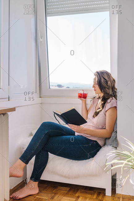 Beautiful 8 months pregnant woman reading a book and relaxing next to the window