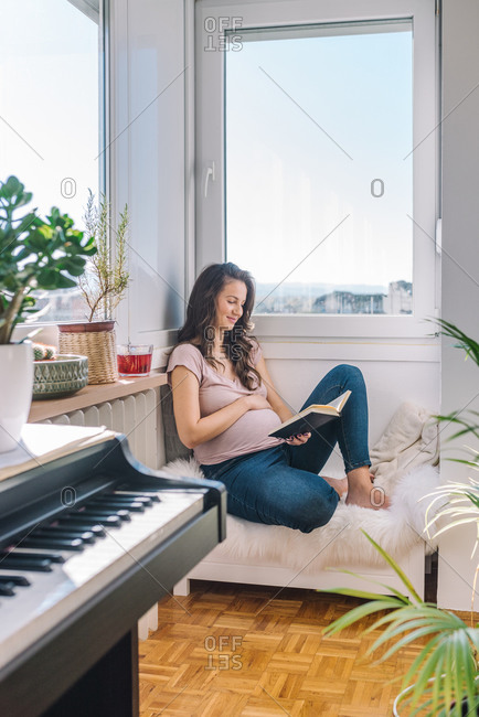Beautiful pregnant woman reading a book and relaxing next to the window
