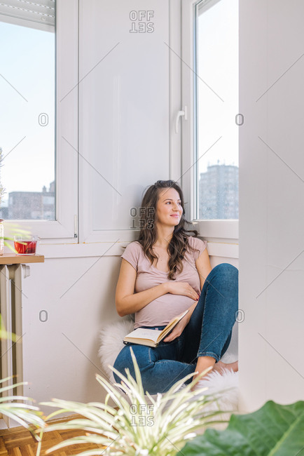 Beautiful pregnant woman looking out window while relaxing and reading a book