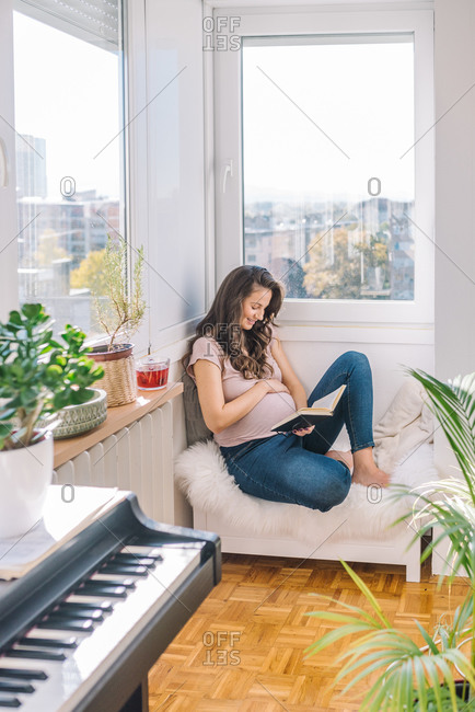 Happy pregnant woman reading a book and relaxing next to the window