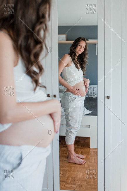 Young pregnant woman looking in mirror while rubbing a stretch mark cream on her belly