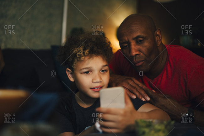 Boy using mobile phone by father in living room at night