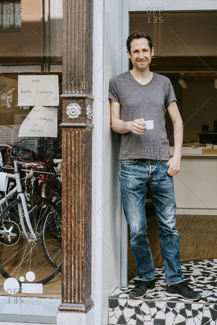 Portrait of smiling male owner with coffee cup standing in doorway of bicycle shop