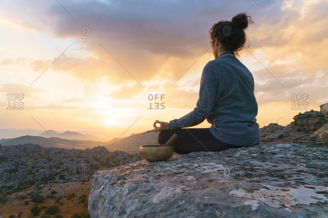 Low angle side view of tranquil female sitting in padmasana position near singing bowl and meditating while relaxing in mountains at sunset time