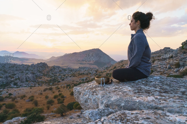 Low angle side view of tranquil female sitting in padmasana position near singing bowl and meditating while relaxing in mountains at sunset time