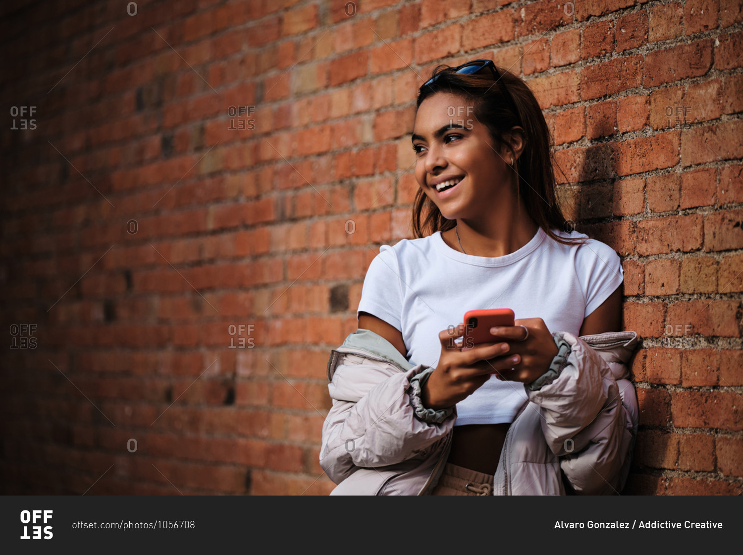 Young cheerful ethnic woman in trendy apparel standing with cellphone near rough wall on street and looking away