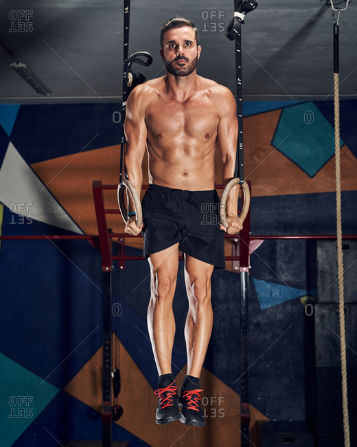 Determined strong young sportsman with naked torso doing pull ups on gymnastic rings during intense workout in gym