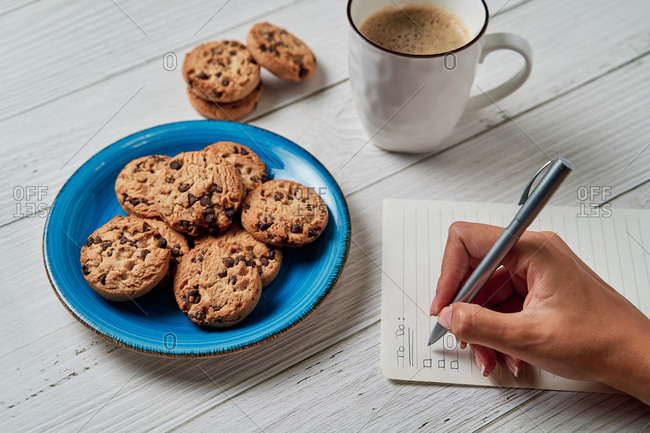 Crop  unrecognizable woman writing to do list on paper while having breakfast with mug of aromatic coffee and crispy cookies