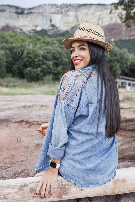 Back view of happy young hipster female traveler in denim jacket and hat sitting on wooden fence against mountains during summer journey in countryside