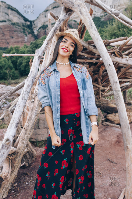 Portrait of ethnic millennial female in bright colorful clothes and hat standing near tent made of dry tree branches against rocky mountains