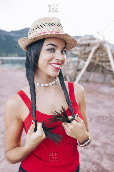 Portrait of ethnic millennial female in bright colorful clothes and hat standing near tent made of dry tree branches against rocky mountains