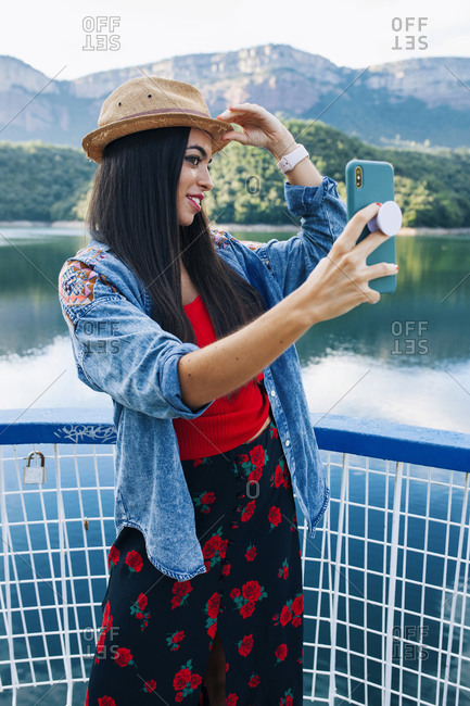 Smiling young female traveler in stylish bright clothing and hat taking selfie on mobile phone while standing near mountain lake during summer holidays