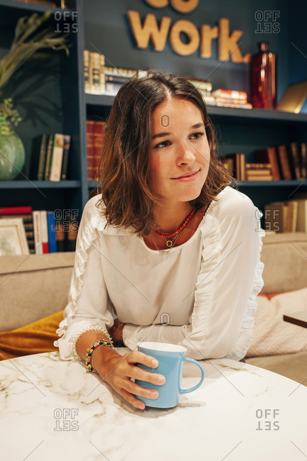 Positive young female in casual outfit sitting at table with cup of coffee and listening attentively to anonymous colleague during meeting in modern workspace