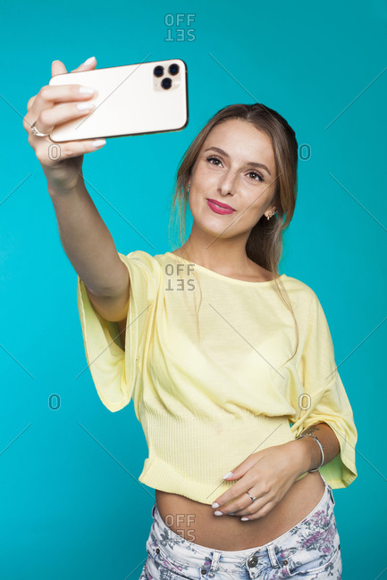 Cheerful coquettish millennial female in casual outfit and with beautiful makeup capturing selfie on mobile phone while standing against bright blue backdrop