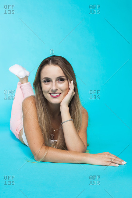 Full length of cheerful dreamy millennial female in casual outfit leaning chin on hand while lying in bright blue studio