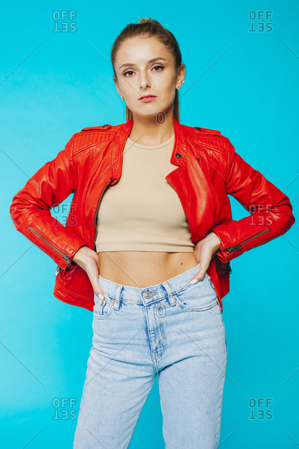 Trendy slim millennial female model in bright red leather jacket and blue jeans standing against vivid blue background