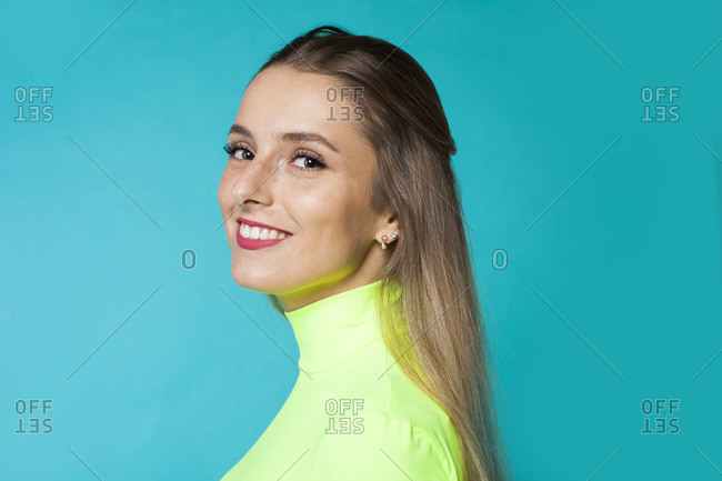 Side view of charming gentle long haired blondie with beautiful makeup wearing bright green turtleneck looking at camera over shoulder against blue background