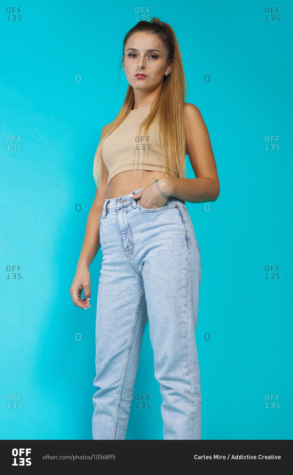 Low angle of confident slim teen female model in cropped top and trendy jeans looking at camera against blue background