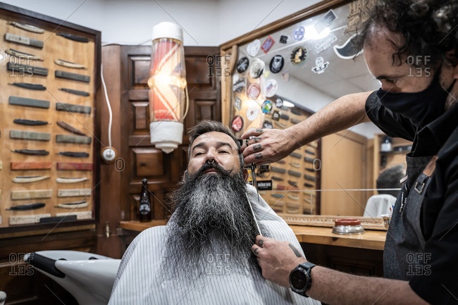 Professional tattooed barber in protective mask with comb and scissors cutting long beard of middle aged gray haired male