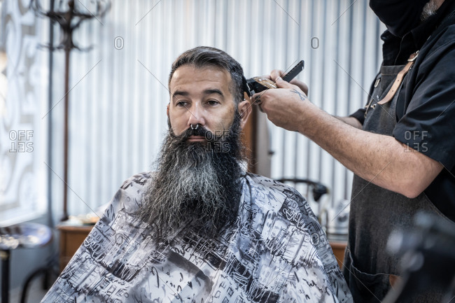 Crop anonymous barber with electric trimmer and comb cutting hair of middle aged hipster male customer with long beard and mustache in modern barbershop