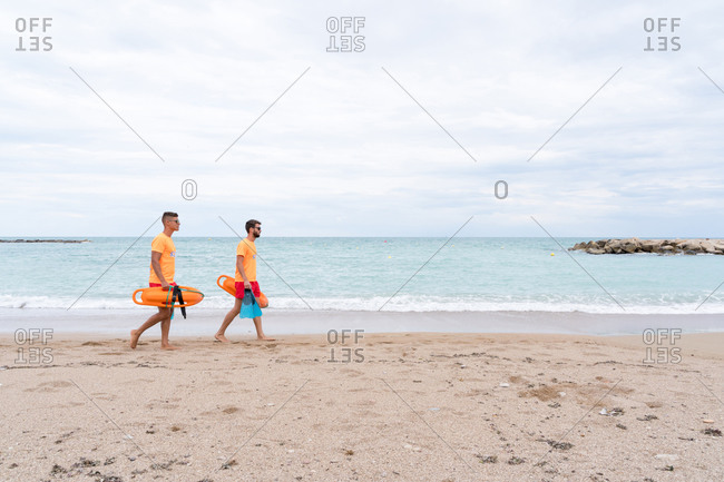 Side view of fit male partners in sunglasses and uniform strolling on sandy ocean coast with torpedo rescue buoys and flippers