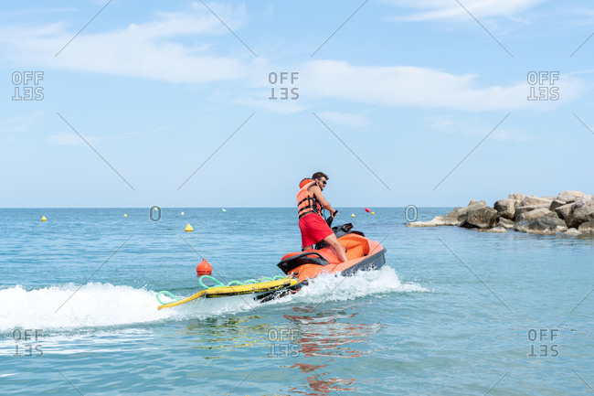 Side view of unrecognizable male worker in life jacket and shorts on bright motor boat on sea under sky