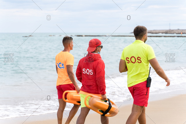 Back view of unrecognizable male friends with torpedo life savers walking on sandy shore near wavy ocean