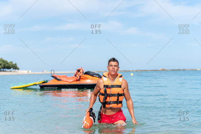 Young lifeguard in bright life vest carrying torpedo rescue buoy on rippled sea near motor boat while looking at camera