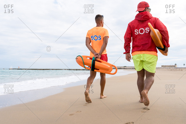 Back view of unrecognizable male friends with torpedo life savers walking on sandy shore near wavy ocean