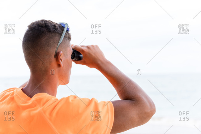 Back view of anonymous male lifeguard in colorful clothes and modern haircut using binoculars while looking at sea