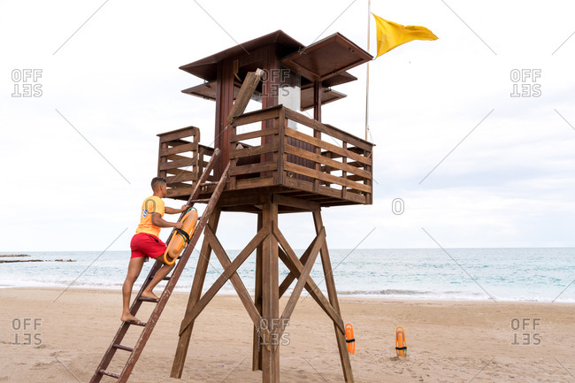 Low angle side view of unrecognizable barefoot lifeguard ascending ladder of wooden tower near sea under cloudy sky