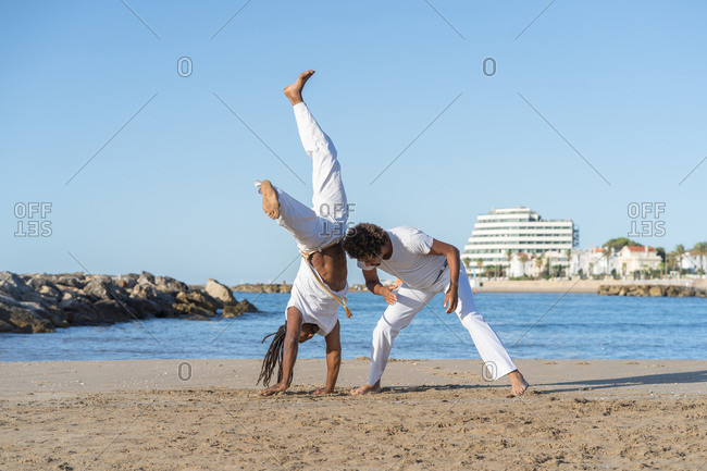 Side view of ethnic african american male partners in white wear fighting while practicing martial art on sea beach